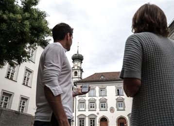 Two people are standing in a historical city view and looking at the tower of a building. The sky is cloudy and the atmosphere is calm.