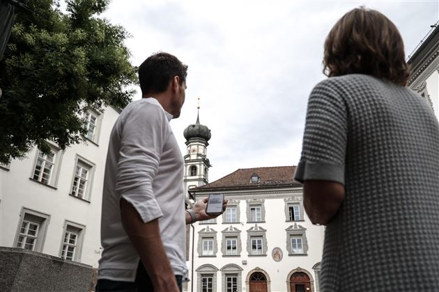 Zwei Personen stehen in einer historischen Stadtansicht und schauen zum Turm eines Gebäudes. Der Himmel ist bewölkt und die Atmosphäre ist ruhig.