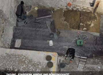 A construction site with working people in a basement room. The floor is uneven and various tools and materials are visible.