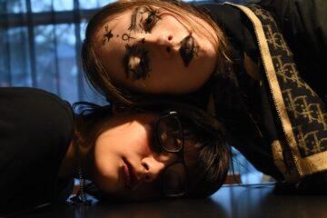 Twee personen liggen naast elkaar op een tafel. Beiden hebben opvallende make-up en een creatieve look.