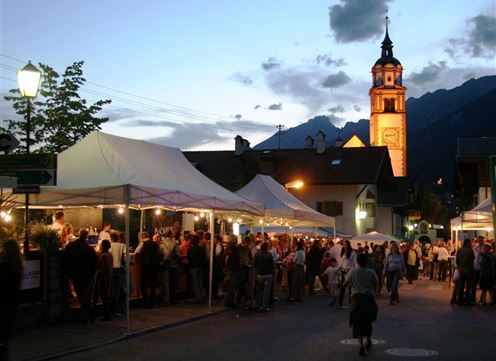 Ein belebter Marktplatz mit Verkaufsständen und vielen Menschen bei Nacht. Im Hintergrund steht ein Kirchturm, der hell beleuchtet ist.
