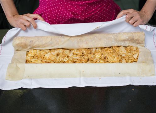 A person is holding a dough with a filling of apple pieces. The dough is being carefully rolled up and is lying on a white cloth.