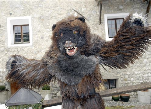 Ein bärähnliches Kostüm mit furchterregendem Gesicht und pelzigen Armen steht vor einem alten Gebäude. Der Hintergrund zeigt Fenster und eine steinerne Wand.
