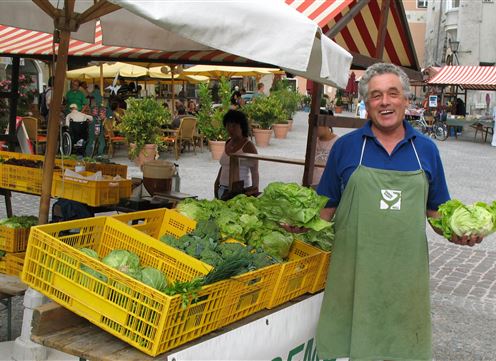 Ein freundlicher Verkäufer steht auf einem Markt und hält frisches Gemüse in den Händen. Im Hintergrund sind weitere Gemüsekisten und Marktbuden sichtbar.