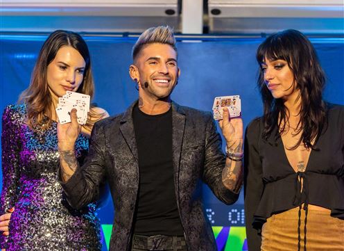 Eine Bühnenaufführung mit einem Magier, der mit Spielkarten posiert. Zwei Frauen stehen neben ihm und schauen interessiert zu.