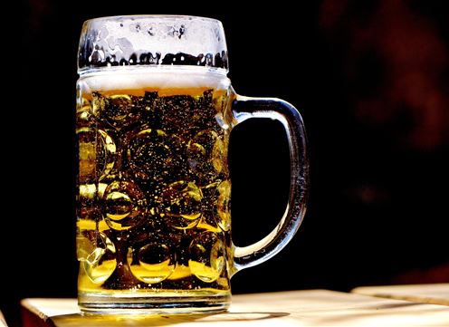 A beer mug filled with frothy, golden liquid is sitting on a table. The glass is clear and the bubbles are visible.