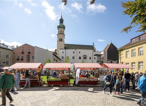 Ein lebendiger Marktplatz mit bunten Verkaufsständen und vielen Menschen. Im Hintergrund ist ein historischer Gebäudeturm zu sehen.