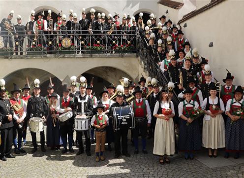 Een grote groep mensen in traditionele kleding staat voor een gebouw. Ze houden instrumenten en bloemen in hun handen.