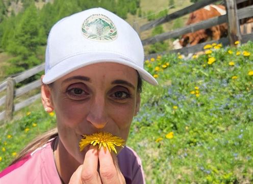 Eine Frau hält eine Löwenzahnblüte vor ihr Gesicht und lächelt. Im Hintergrund sind grüne Wiesen und Kühe zu sehen.