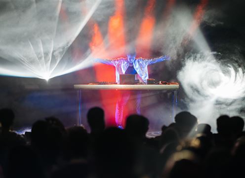 Ein DJ steht hinter einem Mischpult und spielt Musik auf einer Bühne. Bunte Lichter und Nebel sorgen für eine lebhafte Atmosphäre.