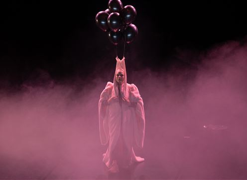 Eine Person in einem auffälligen Kostüm hält einen Strauß schwarzer Ballons. Der Hintergrund ist von rosa Nebel umgeben.