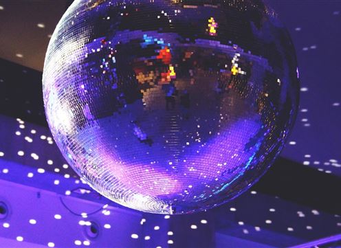 Eine glänzende Diskokugel hängt an der Decke und reflektiert das Licht. Der Raum ist in sanften Lila- und Blautönen beleuchtet.