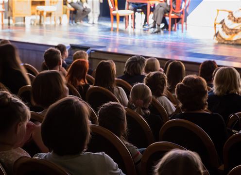 A crowd of spectators is sitting in a theater and looking at the stage. The atmosphere is both relaxed and tense.
