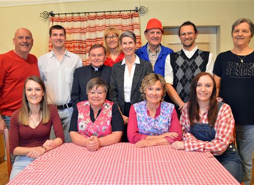 A group of people is standing and sitting at a table with a checkered tablecloth. They are laughing and seem to be having a good time together.