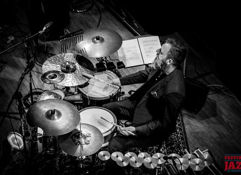A drummer plays on his drum during a jazz festival. Sheet music is visible in the background.