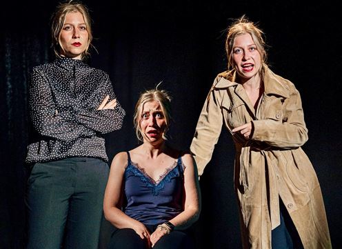 Three actresses are standing on stage. One is sitting in the middle, while the other two are staring at her, creating a tense atmosphere.
