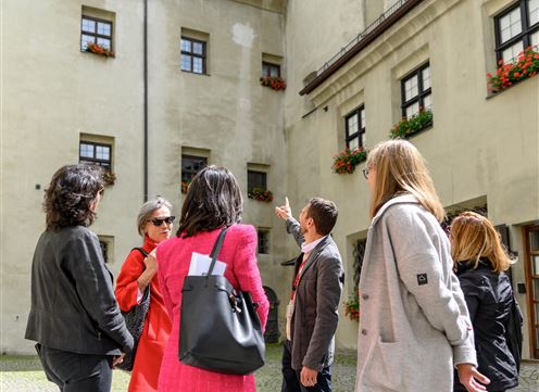 Eine Gruppe von Menschen steht in einem Innenhof und hört einem Führer zu. Alte Gebäude mit Fenstern und Pflanzen sind im Hintergrund zu sehen.