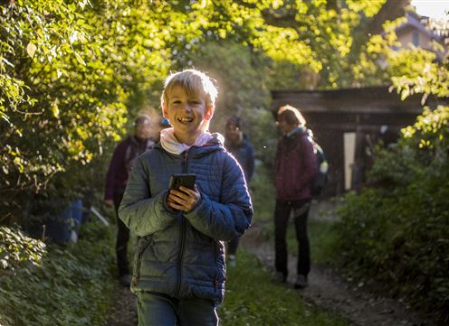 Een vrolijke jongen met een smartphone loopt op een pad, omgeven door bomen. Op de achtergrond zijn meer mensen te zien die ook aan het wandelen zijn.