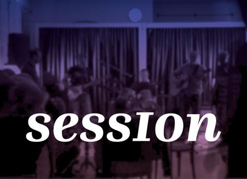 Eine Musiksession mit mehreren Musikern auf einer Bühne. Die Atmosphäre ist lebhaft und die Zuschauer sind aufmerksam.