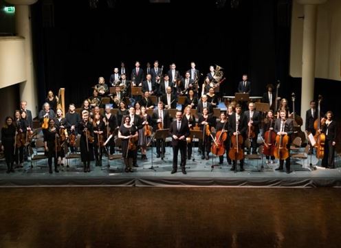 Ein ganzes Orchester steht auf der Bühne in eleganter Kleidung. Die Musikerinnen und Musiker halten ihre Instrumente und blicken in die Richtung des Zuschauers.