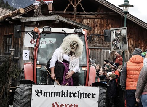 Ein Traktor mit verkleideten Personen fährt durch eine Menschenmenge. Die Darstellung zeigt eine regionale Feier mit traditionellem Fokus auf Hexen.