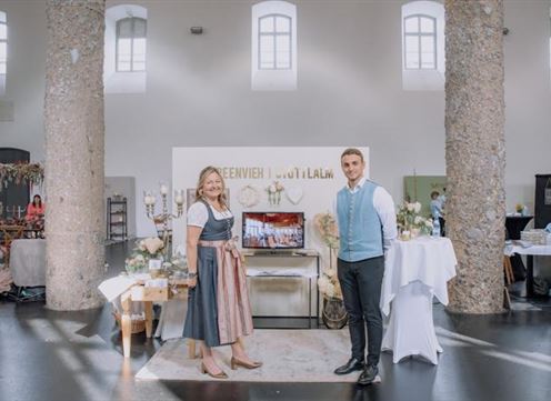 A festive room with two people smiling warmly at the camera. In the background, decorative elements and tables with flower arrangements can be seen.