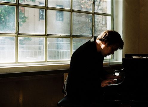 A musician is playing the piano in a bright room. Sunlight streams through a large window.