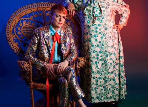 Ein Paar Personen posiert in einem stilvollen, bunten Setting. Die eine Person sitzt auf einem Stuhl und trägt einen glitzernden Anzug, während die andere in einem floralen Kleid steht.