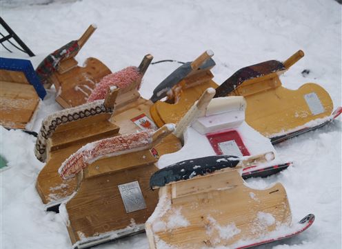 A collection of sleds lies in the snow. The sleds are made of wood and are in various colors.