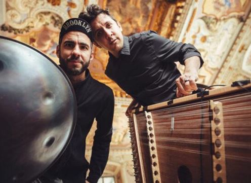 Twee muzikanten staan in een mooie ruimte met een fraai plafond. De man links speelt een handpan, terwijl de ander een ander instrument presenteert.