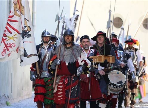 Eine Gruppe von historischen Soldaten marschiert durch eine schmale Gasse. Sie tragen Rüstungen und halten Flaggen sowie Musikinstrumente.