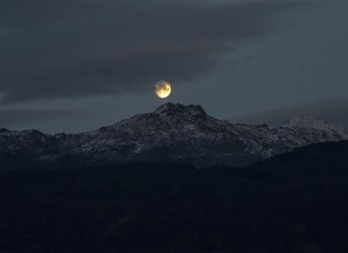 Ein schöner Vollmond, der über schneebedeckten Bergen aufgeht. Der Himmel ist bewölkt und verleiht der Szene eine mystische Atmosphäre.