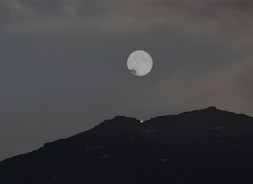 Een grote, heldere maan staat boven een donkere berg. De lucht is licht bewolkt en heeft een mystieke uitstraling.