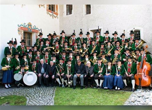 Eine große Blasmusikkapelle in traditionellen Trachten steht im Freien. Die Musiker tragen grüne Jacken und schwarze Hüte, und einige Instrumente sind gut sichtbar.