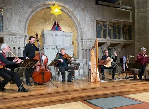 A musical performance in a festive space. Six musicians play various instruments in front of a decorative backdrop with a statue.