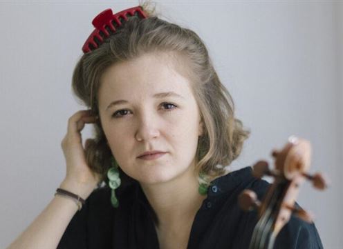 A young woman with curly hair and a red hairpin is looking directly at the camera. Next to her is a musical instrument, possibly a violin.