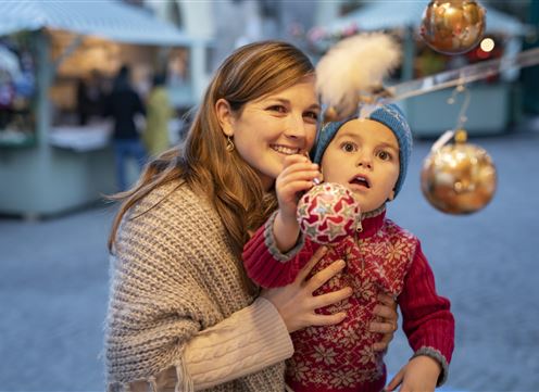 A smiling mother holds her child in her arms. In the background, Christmas decorations and stands can be seen.