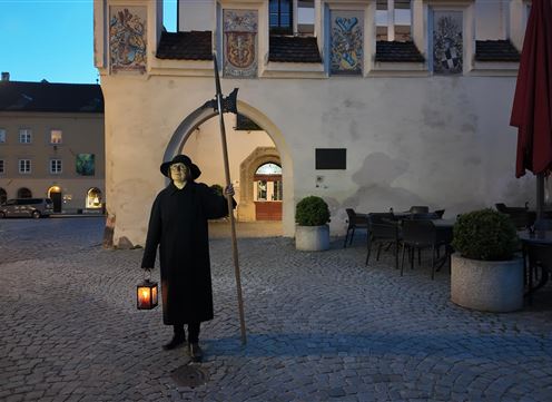 Eine Person in schwarzer Kleidung steht mit einer Laterne und einem Stock in einem historischen Platz. Die Szenerie ist abends, mit einem ruhigen, stimmungsvolle Licht.