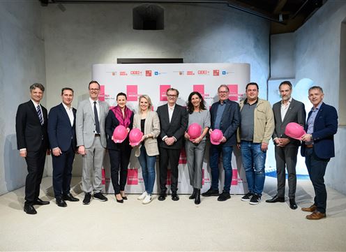 Eine Gruppe von Personen steht zusammen in einem Raum mit einem Banner im Hintergrund. Alle tragen rosa Helme und lächeln freundlich in die Kamera.