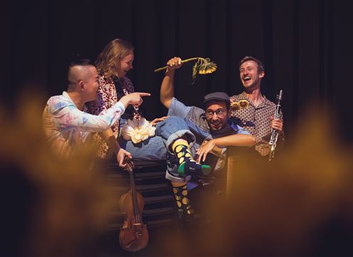 A cheerful music group poses on stage. They are dressed in colorful clothing and laugh while holding various instruments.