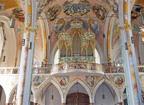 A magnificent church interior with a large organ in the foreground. The walls are artistically designed with frescoes and decorative elements.