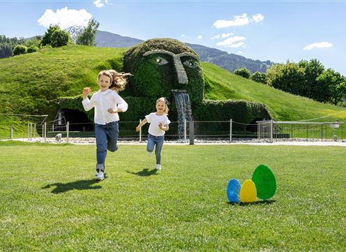 Twee meisjes lopen vrolijk over een groene weide. Op de achtergrond is een grote, mooie sculptuur van hagen te zien.