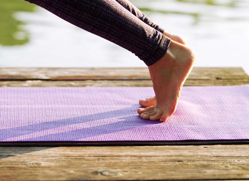 A person is standing on a purple yoga mat at the edge of a wooden walkway. In the background, water can be seen.