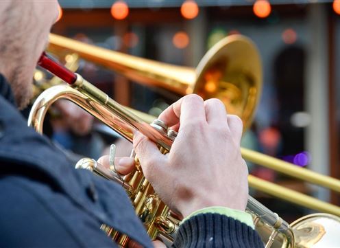 Ein Musiker spielt Trompete und hält das Instrument mit einer Hand. Die Umgebung ist lebhaft mit verschwommenen Menschen im Hintergrund.