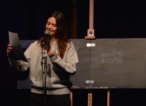 A man is standing on a stage and speaking into a microphone. On a table next to him, drinks and notes are visible.