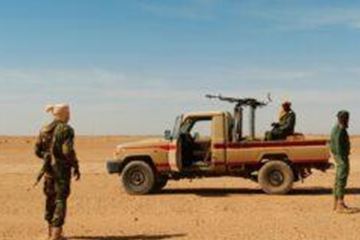 Een groep soldaten staat in een schrale woestijnlandschap. Een gepantserd voertuig is op de achtergrond te zien.