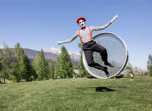 Ein Clown in gestreifter Kleidung springt fröhlich auf einer großen, runden Plattform. Im Hintergrund sieht man grüne Bäume und Berge.