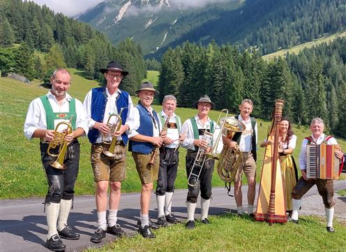 A group of musicians in traditional attire stands in a green landscape. In the background, mountains and forests can be seen.
