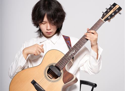 Ein junger Musiker spielt Gitarre und konzentriert sich auf die Saiten. Er trägt ein weißes Hemd und sitzt auf einem Stuhl.