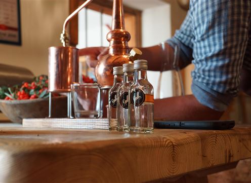 Eine Person bereitet mit einer kupfernen Anlage Likör in einer gemütlichen Küche vor. Auf dem Tisch stehen mehrere kleine Flaschen bereit.
